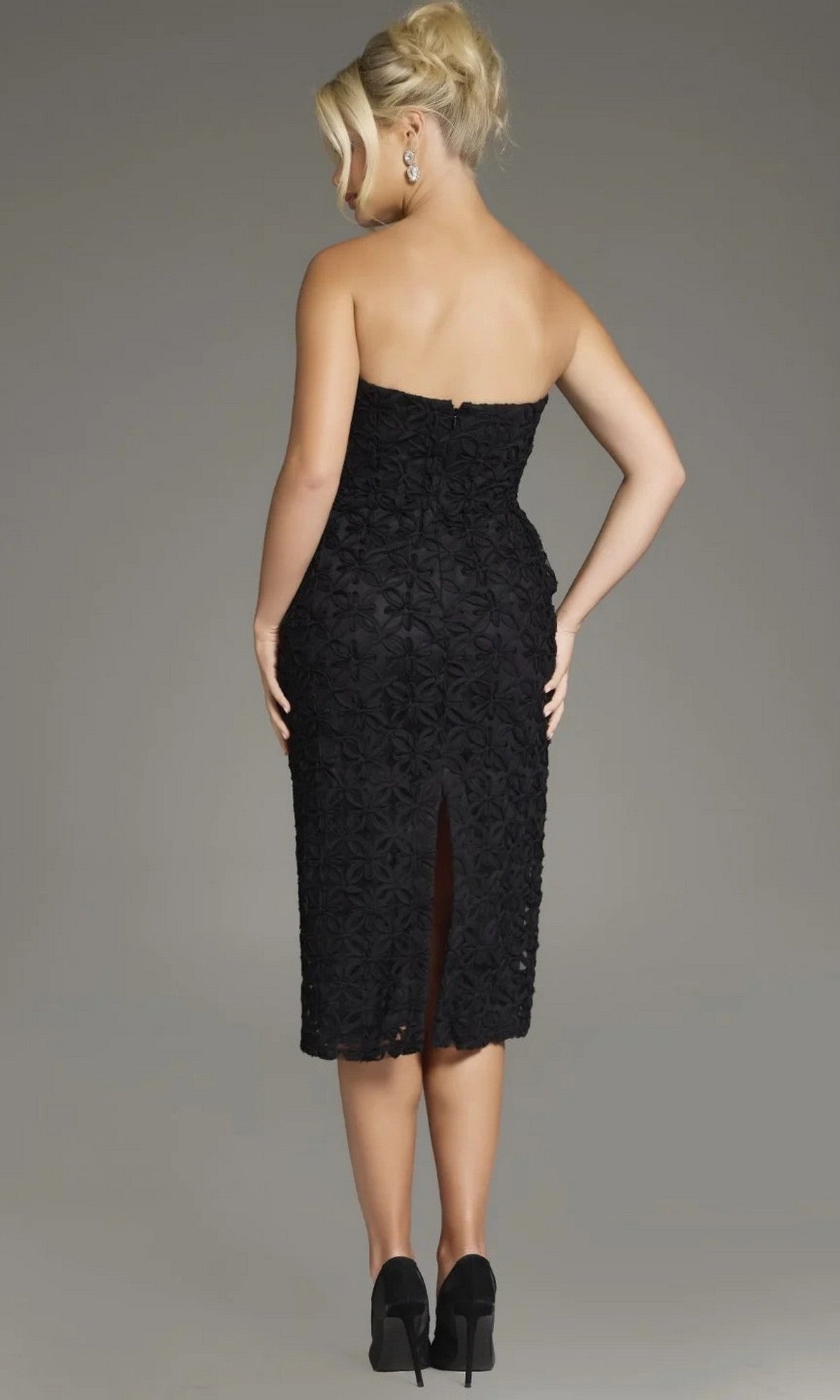 A woman with blonde hair in an updo is wearing the Jovani 39076 Midi Wedding Guest Dress, a strapless black lace formal dress, along with black heels, standing with her back to the camera against a plain background.